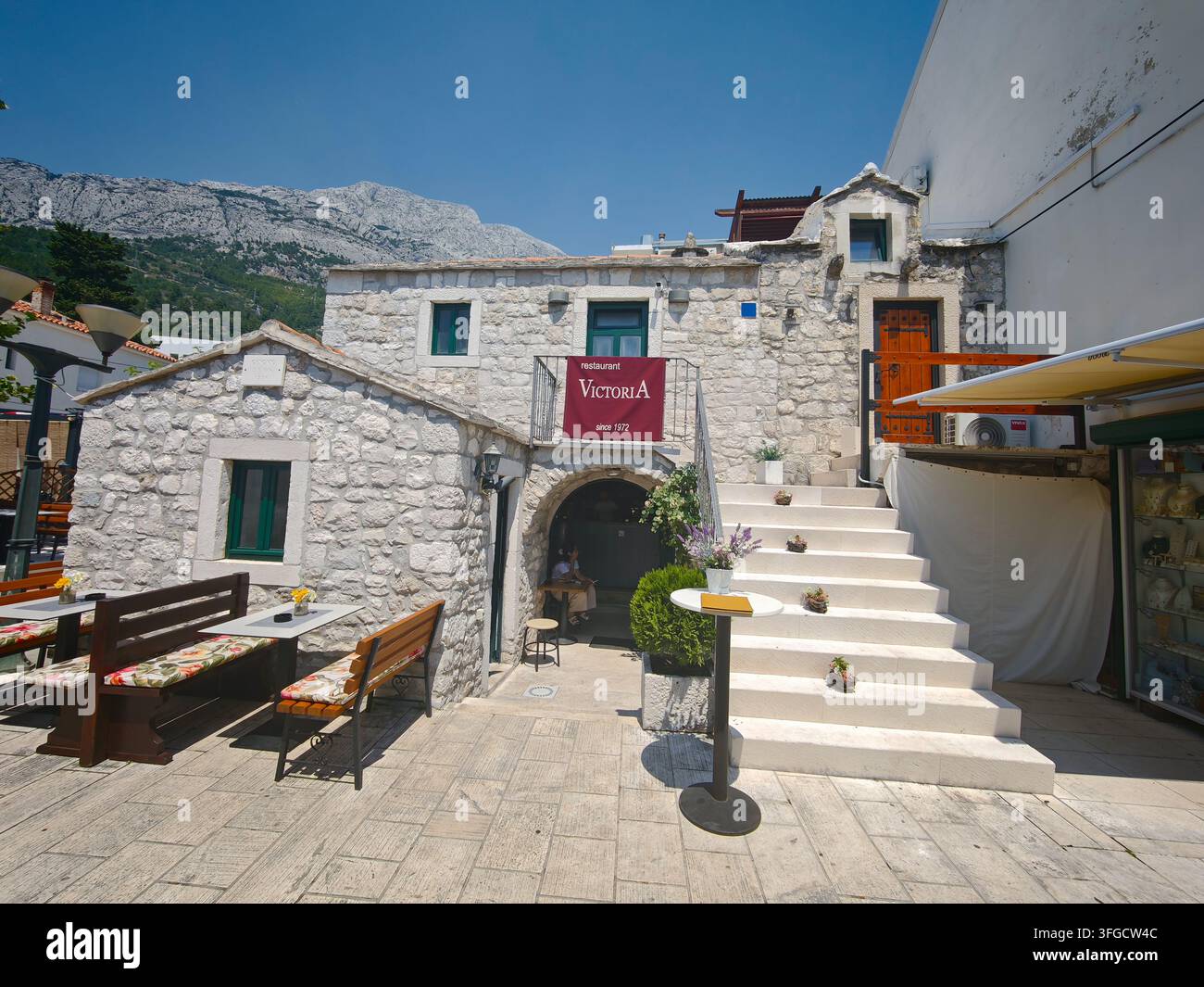 Une vue panoramique de l'extérieur du restaurant traditionnel en pierre avec des places de siège en plein air, des marches en pierre blanche et un majestueux fond montagneux à Baska Voda. - Image de stock capturée avec un smartphone