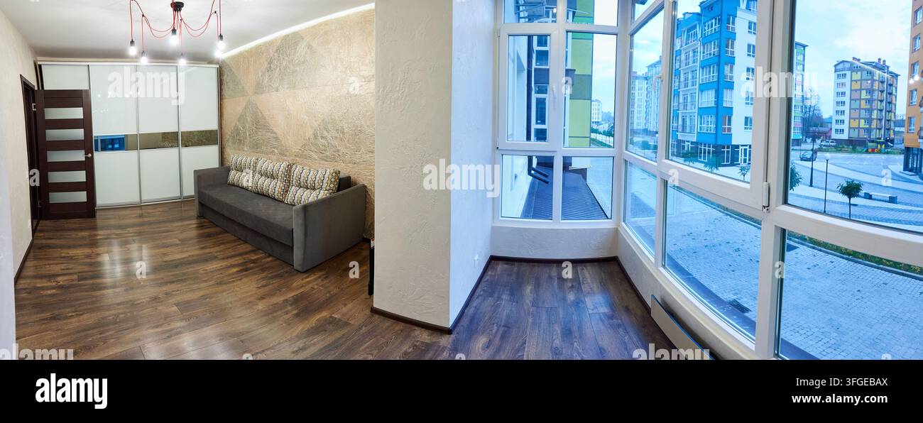 Intérieur d'appartement moderne avec canapé gris, armoire coulissante et fenêtres panoramiques donnant sur des bâtiments résidentiels. Espace de vie élégant avec sol en bois et concept de design contemporain Banque D'Images