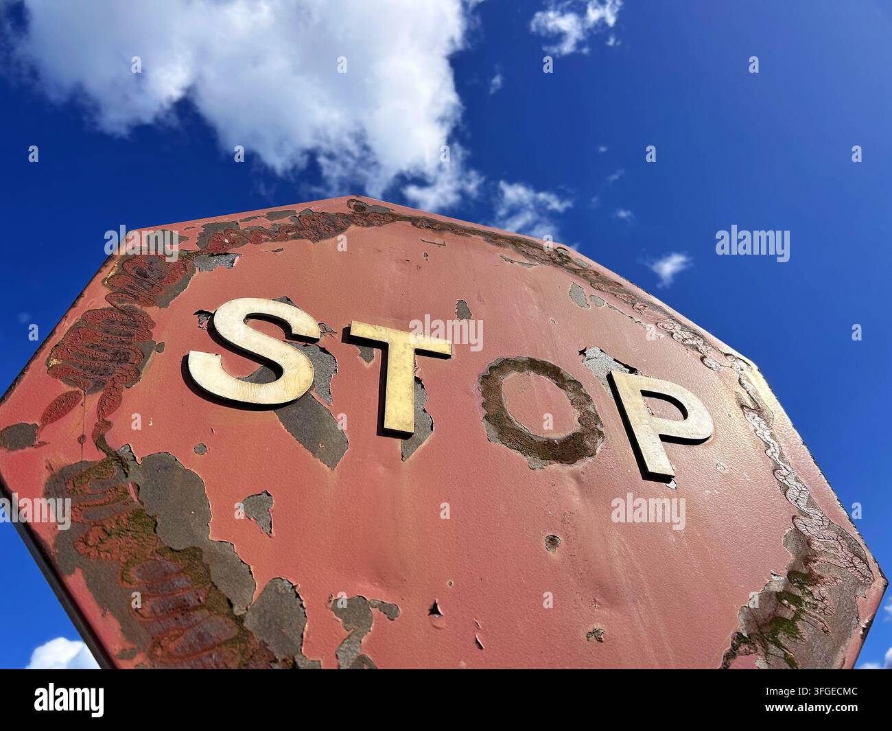 Gros plan d'un vieux panneau STOP érodé avec de la rouille, de la peinture rouge écaillée et des lettres blanches en relief contre un ciel bleu clair Banque D'Images
