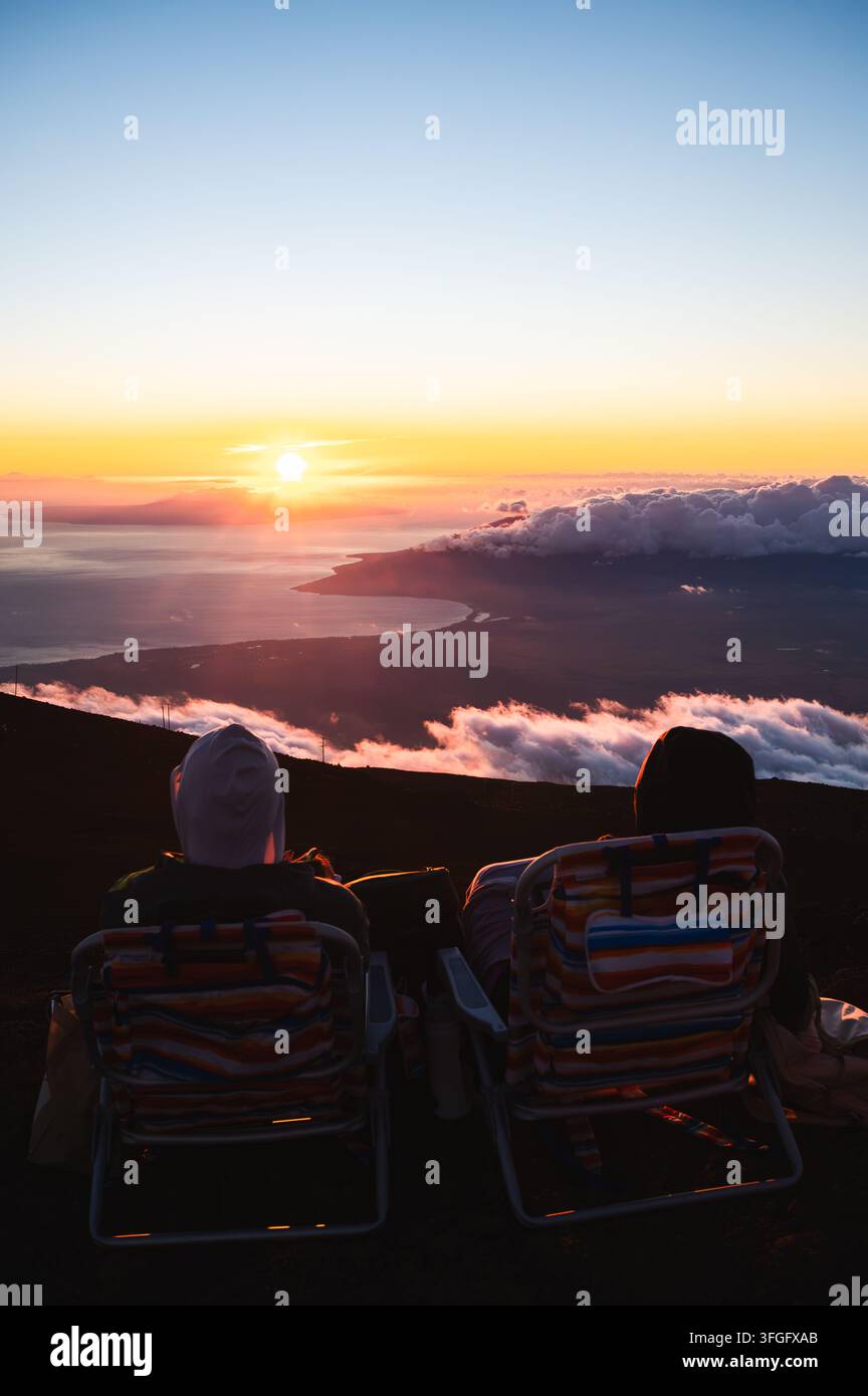 Couple se relaxant dans des chaises longues et regardant le coucher de soleil au-dessus des nuages depuis le sommet du volcan Haleakala, Maui, Hawaï, États-Unis Banque D'Images