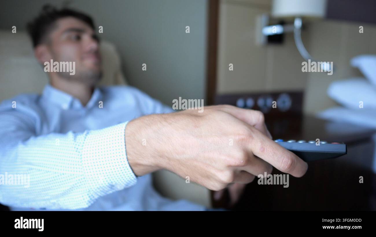 Focus sur la main masculine du jeune homme d'affaires dans les canaux de commutation de fauteuil sur la télécommande de télévision à la chambre d'hôtel moderne. Détente cosy lors d'un voyage d'affaires Banque D'Images