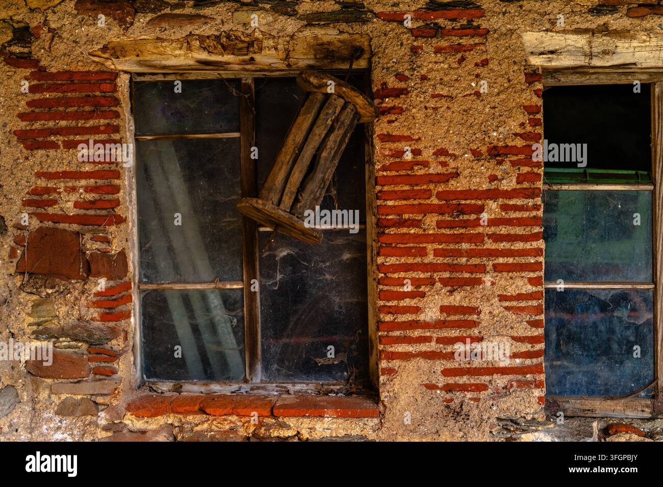Deux vieilles fenêtres avec des cadres en bois encastrées dans un mur de briques et de plâtre délabré, avec une chaise en bois patinée visible à travers l'une des ouvertures. Banque D'Images