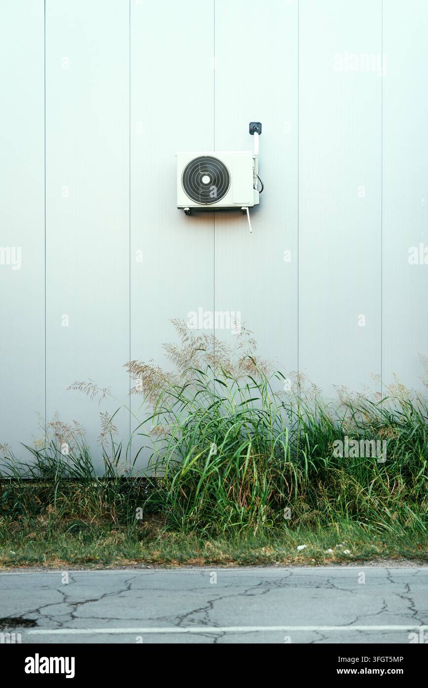 Unité AC extérieure blanche montée sur une façade de bâtiment moderne minimaliste au-dessus d'une haute herbe. Concept de contrôle climatique, consommation d'énergie, technologie de refroidissement Banque D'Images