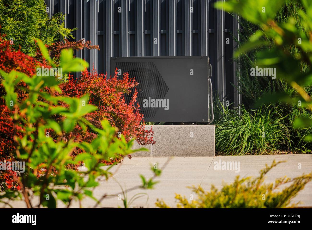 Unité de pompe à chaleur extérieure installée près d'un bâtiment moderne, entourée de plantes ornementales et d'arbustes colorés par une journée ensoleillée Banque D'Images