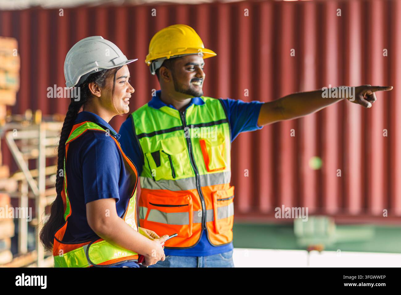 Ouvriers d'entrepôt formant une jeune travailleuse, main pointant souriante, portant casque et gilet de sécurité, équipe de conteneurs d'expédition. Masculin Féminin Superviseur Logistique Banque D'Images