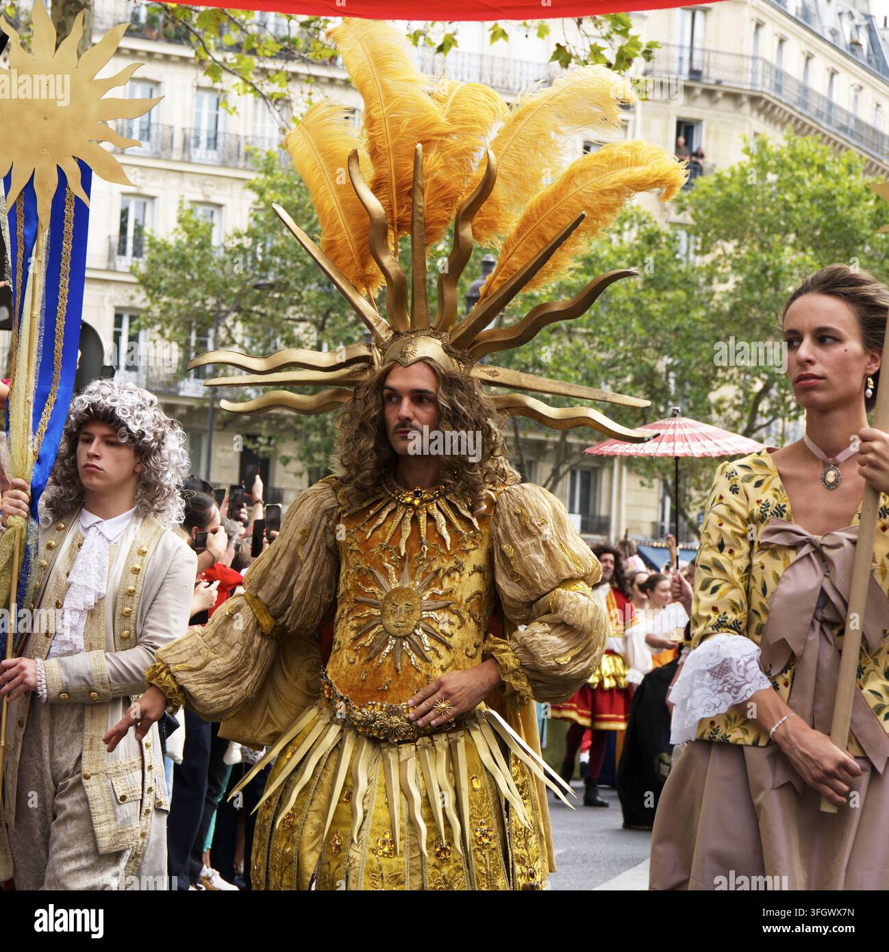 Un artiste vêtu d'un costume orné d'or du Roi Soleil avec un couvre-chef en forme de soleil à plumes, défilant aux côtés d'assistants en tenue de cour baroque du XVIIe siècle Banque D'Images