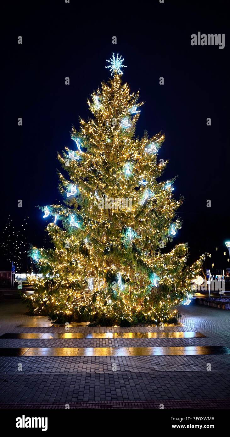 Sapın de Noël illuminé décoré de guirlandes lumineuses chaudes et d’ornements d’oiseaux blancs sur une place publique à Anykščiai, Lituanie. - Image de stock capturée avec un smartphone
