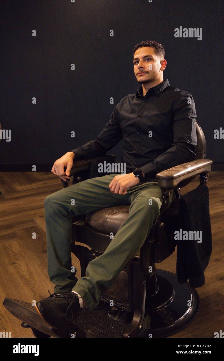 Homme adulte assis dans un fauteuil de coiffeur vintage dans un coin du studio, portant une chemise noire et une veste sombre Banque D'Images