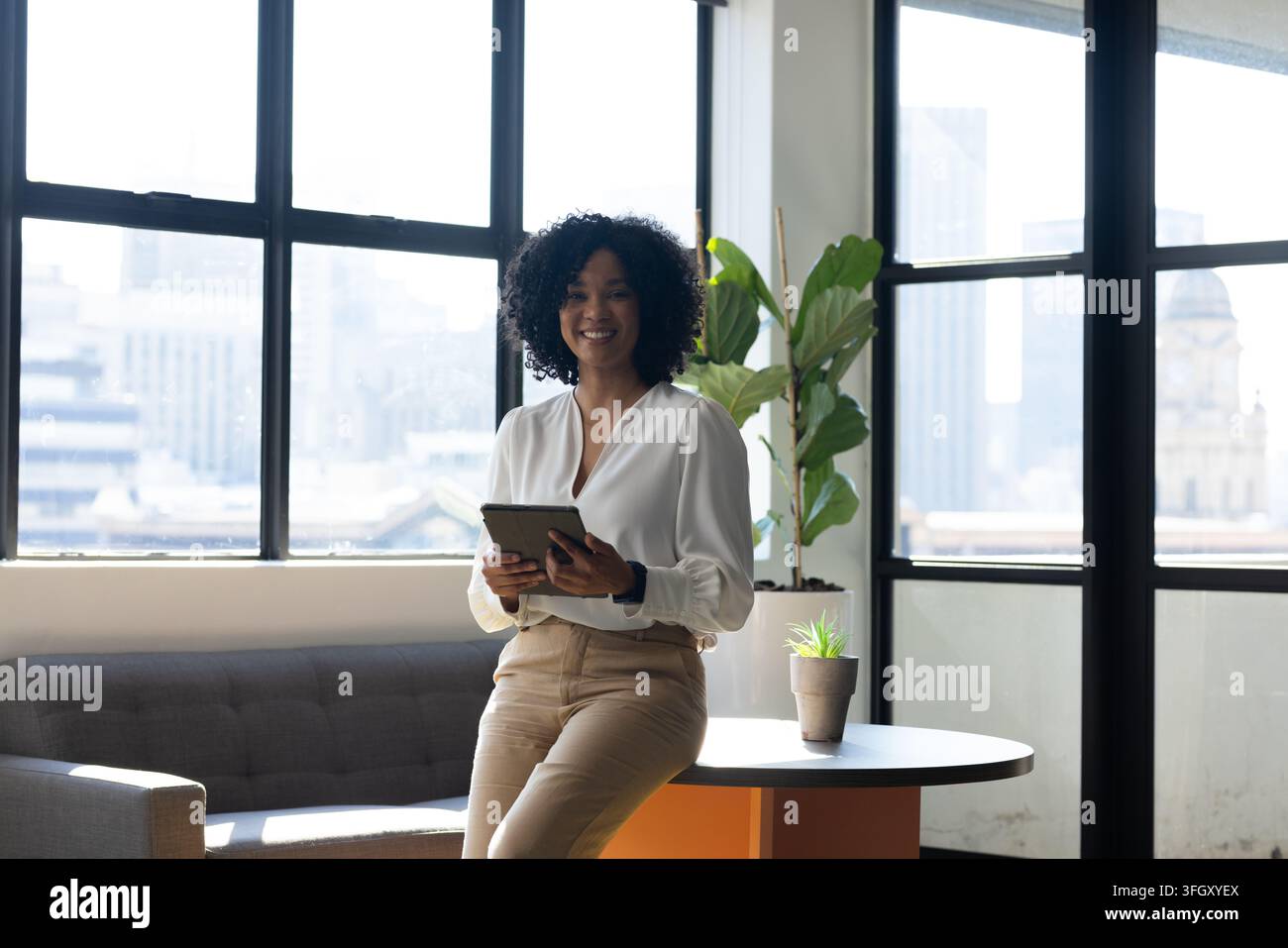 Femme afro-américaine appuyée à une table dans un bureau tenant une tablette, portant un chemisier blanc et un pantalon beige Banque D'Images