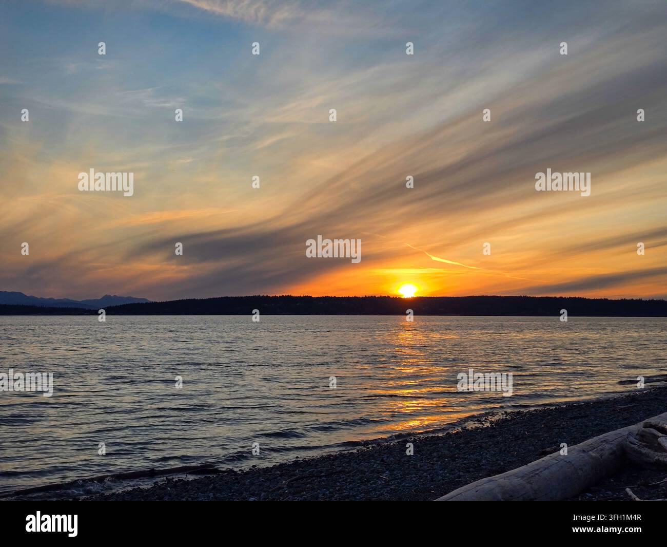 Un coucher de soleil doré brille au-dessus d'une ligne d'horizon lointaine alors que des nuages striés dramatiques remplissent le ciel et que la lumière chaude se reflète sur l'eau ondulante vers une rive rocheuse Banque D'Images