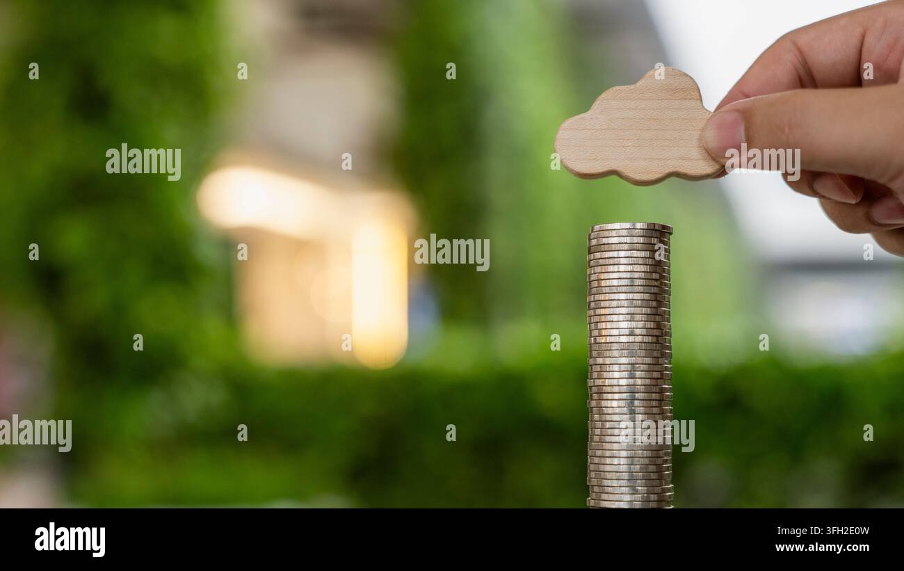 Concept de coûts d'investissement en cloud computing et de financement des technologies numériques avec une main tenant une icône de nuage en bois au-dessus de pièces empilées dans un bureau moderne Banque D'Images