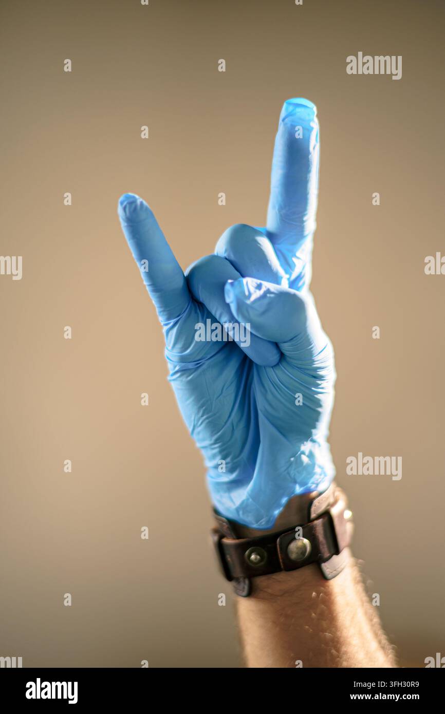 Gros plan d'une main gantée bleue jetable faisant un geste rock and roll ou cornes contre un fond chaud et uni. Banque D'Images