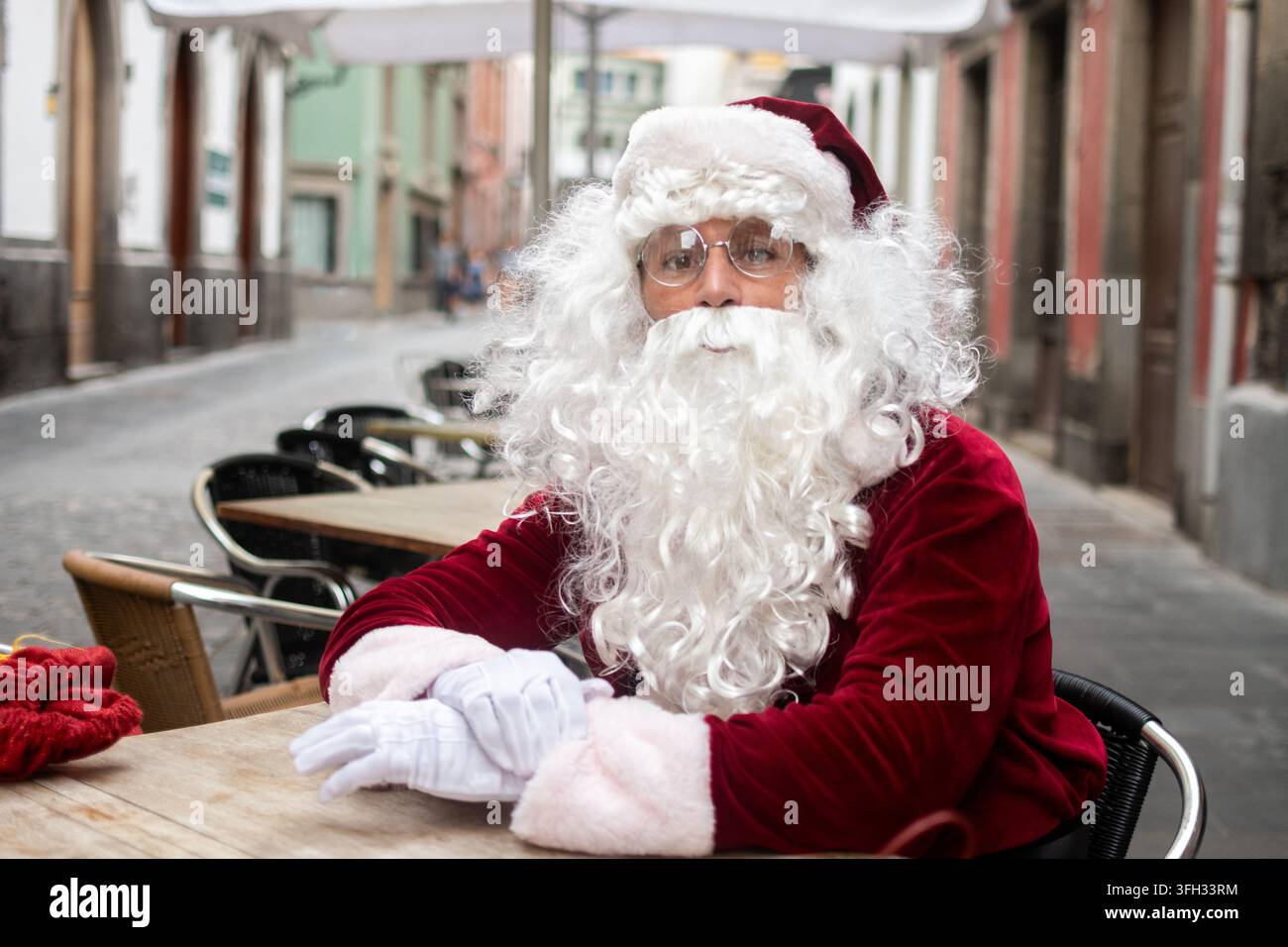 Le Père Noël vêtu d'un costume traditionnel rouge et blanc assis à une table de café en plein air dans une rue urbaine calme pendant la saison de Noël. Banque D'Images