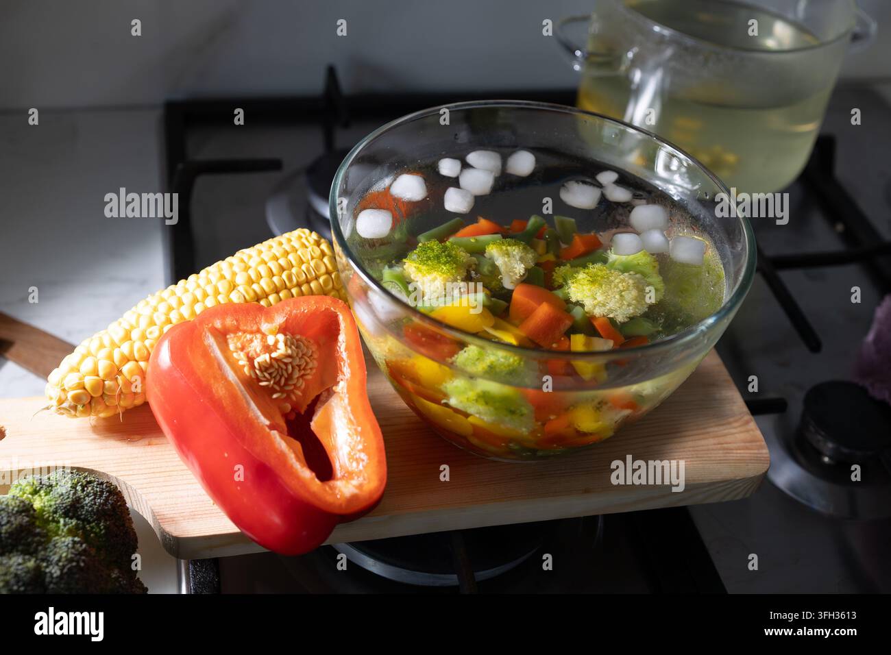 Un bol en verre sur un comptoir de cuisine contient des légumes blanchis refroidissant dans un bain d'eau glacée, avec des glaçons et une cuisinière visibles en arrière-plan de la cuisine Banque D'Images