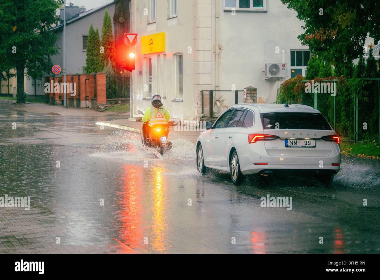 VALMIERA, LETTONIE - 1er SEPTEMBRE 2026 : Apprenti motard roule sous une forte pluie aux côtés de la circulation sur une rue de ville inondée Banque D'Images