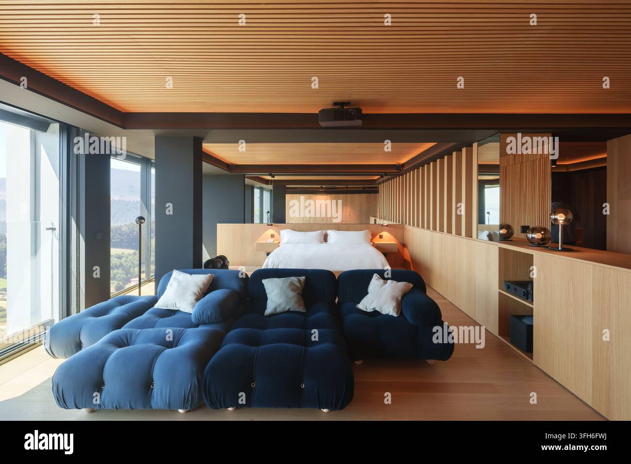 Chambre côtière moderne et chaleureuse avec intérieur en bois, canapé bleu foncé capitonné, lit plateforme, fenêtres panoramiques et vues paisibles sur le paysage montagneux Banque D'Images
