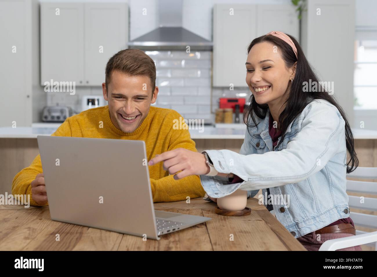 Couple assis dans la cuisine utilisant un ordinateur portable argenté, homme en pull jaune, femme avec bandeau rose pointe Banque D'Images
