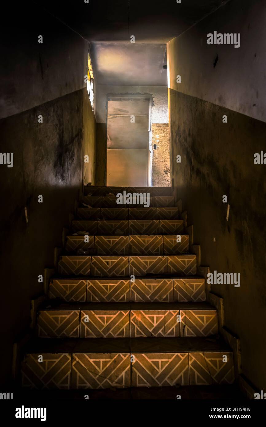 Escalier abandonné et effrayant dans un vieux bâtiment avec des carreaux à motifs géométriques sur les marches, menant à une porte éclairée mystérieuse au Brésil. Banque D'Images
