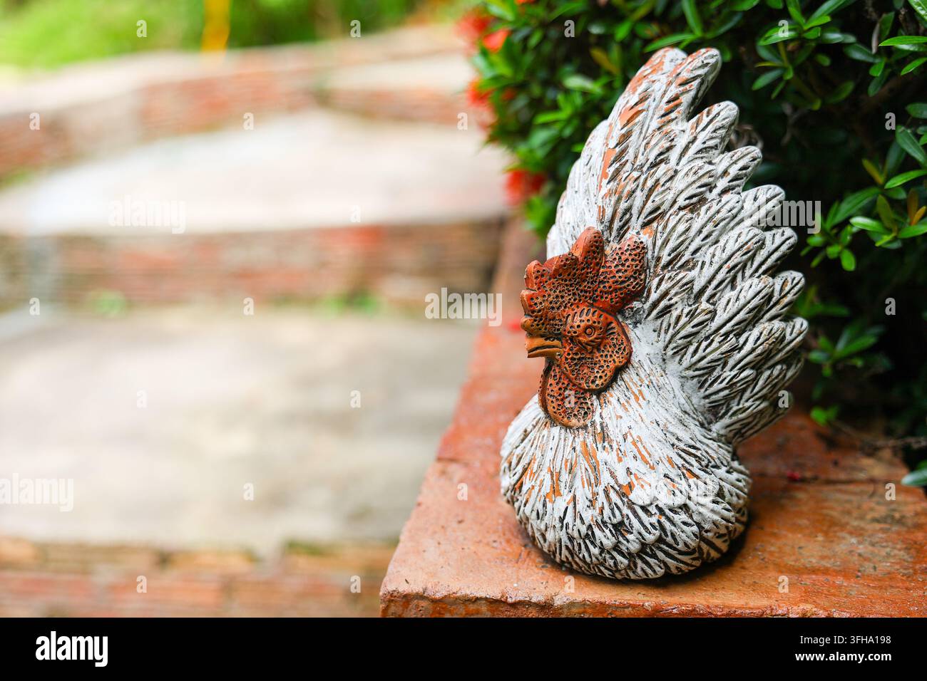 Une figurine décorative de coq avec texture de plumes à côté d'un arbuste feuillu sur un rebord de brique créant une scène de décoration de jardin rustique Banque D'Images