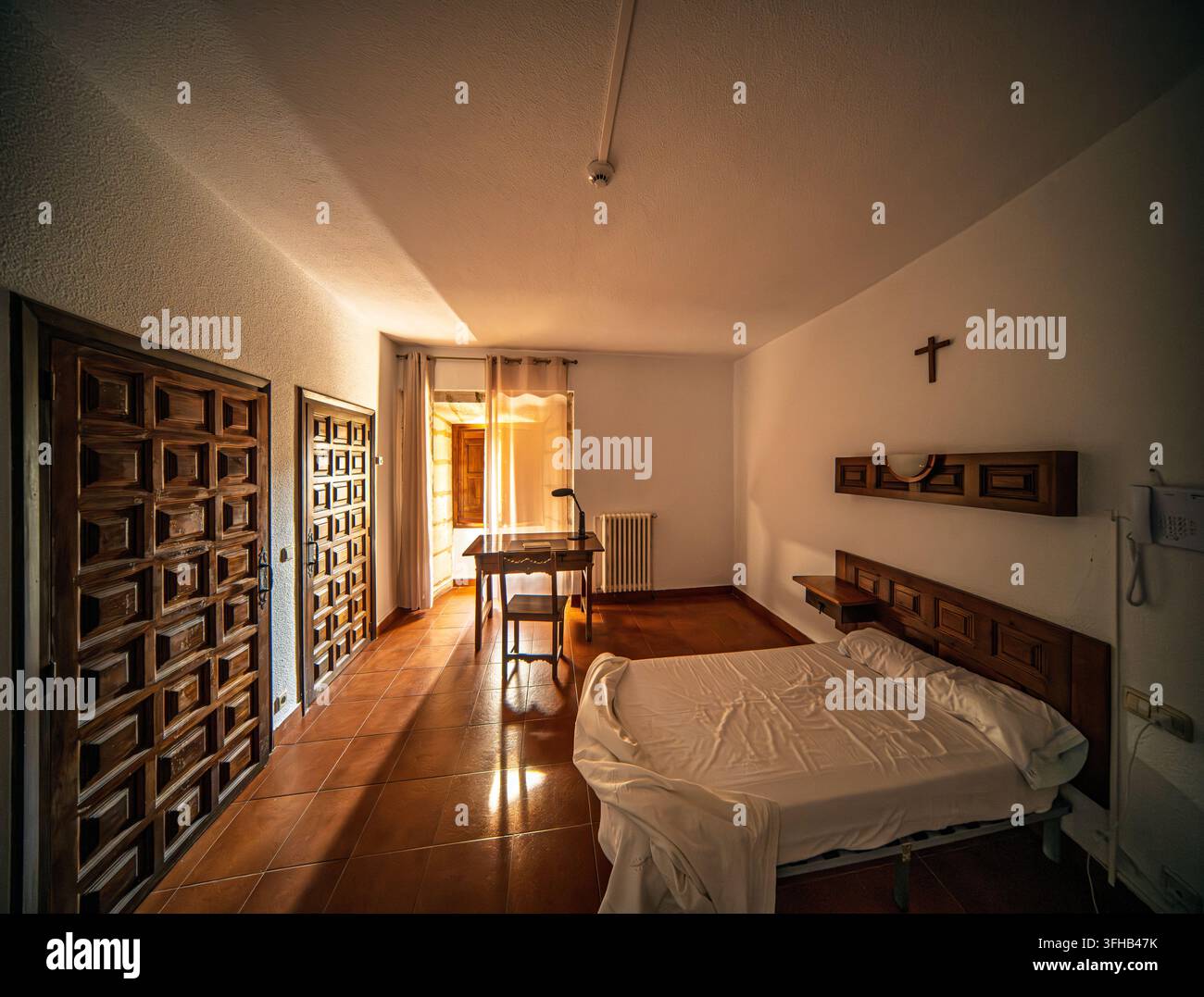 Chambre de gîte austère baignée de soleil avec un lit, un bureau et une croix au mur à l'abbaye de Santo Domingo de Silos, Burgos, Castille-et-León, Espagne. Banque D'Images