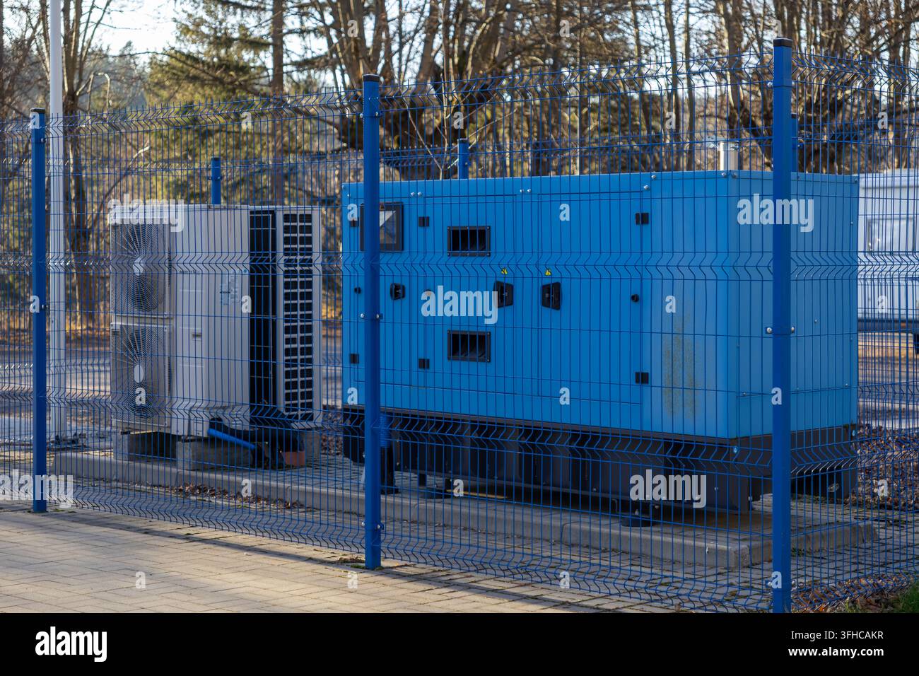 Un grand générateur industriel bleu et une unité de climatisation blanche se dressent derrière une clôture métallique bleue de protection, fournissant une alimentation de secours et un climatage critiques Banque D'Images