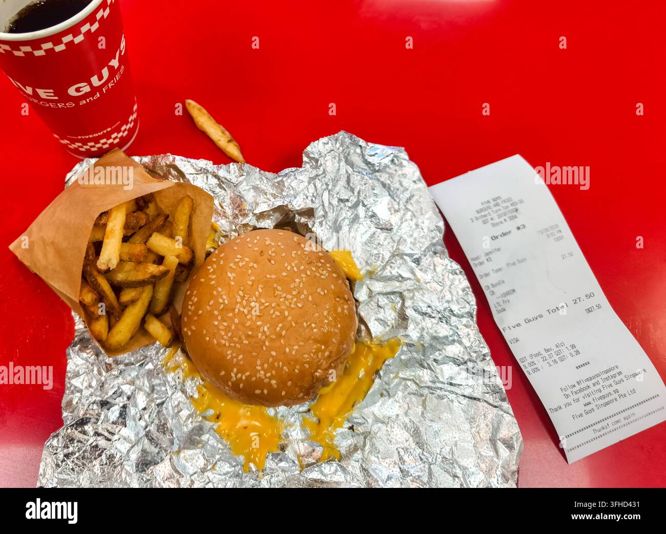 Repas de fast-food de Five Guys, comprenant un cheeseburger enveloppé dans du papier d'aluminium, un sac en papier brun rempli de frites fraîchement coupées, un gobelet à boisson et un reçu sur une table rouge Banque D'Images