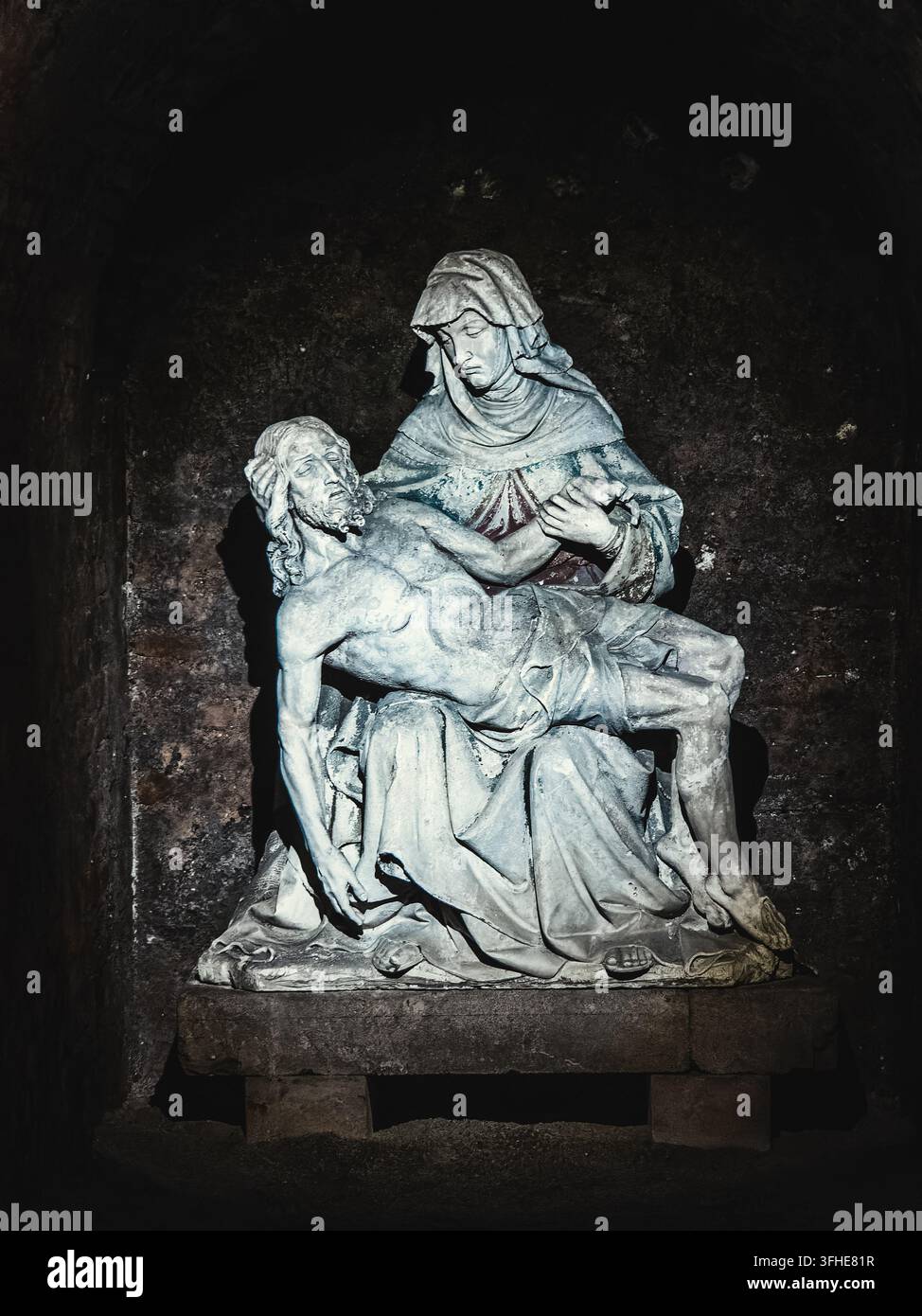 Une sculpture en pierre usée de Pietà représente une mère en deuil berçant une figure sans vie, dramatiquement éclairée dans une alcôve sombre de pierre vieillie. Banque D'Images