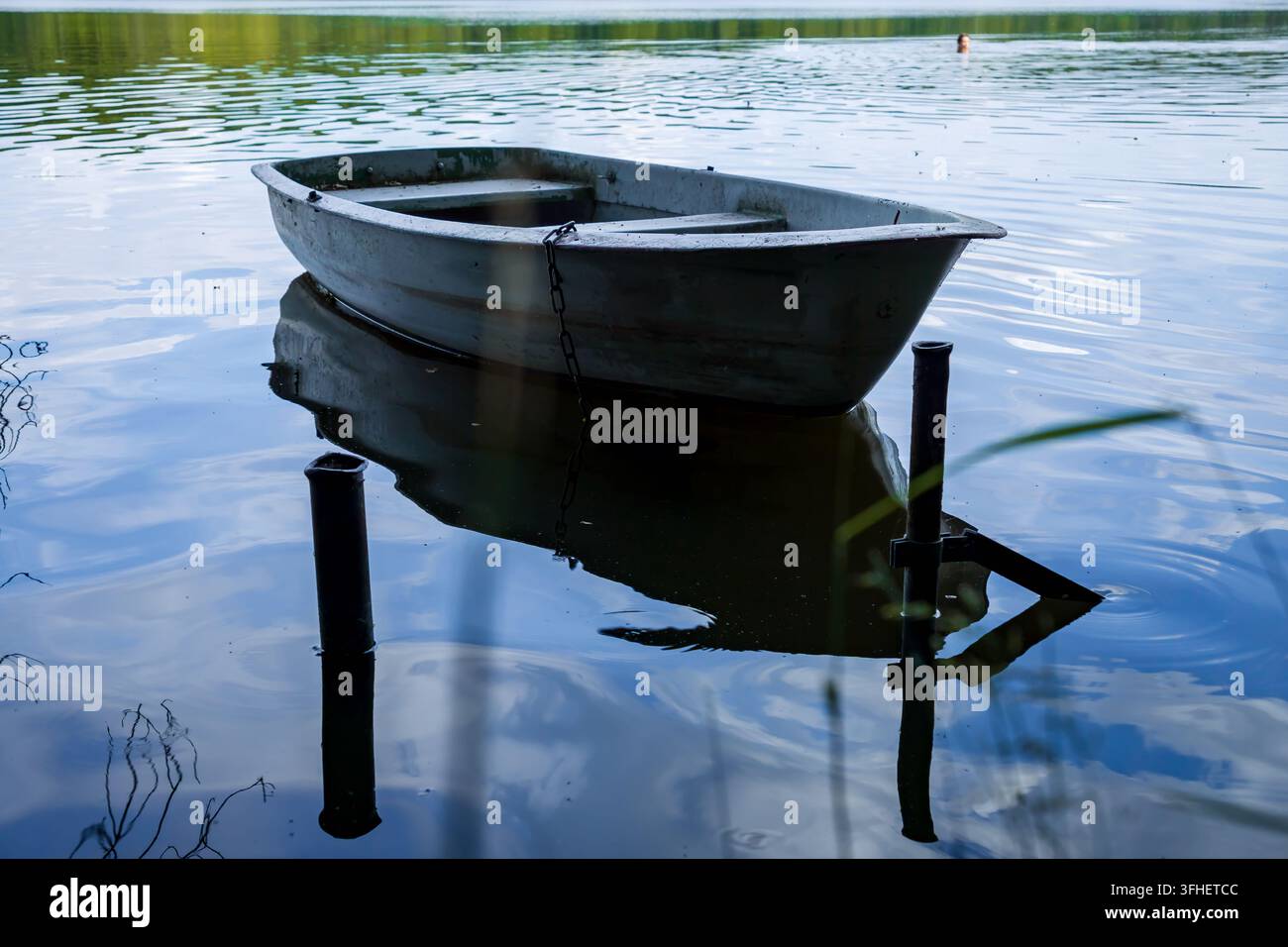 Une barque métallique vide amarrée sur un lac forestier calme, avec des reflets sur l'eau et un nageur lointain flou en arrière-plan. Été paisible Banque D'Images