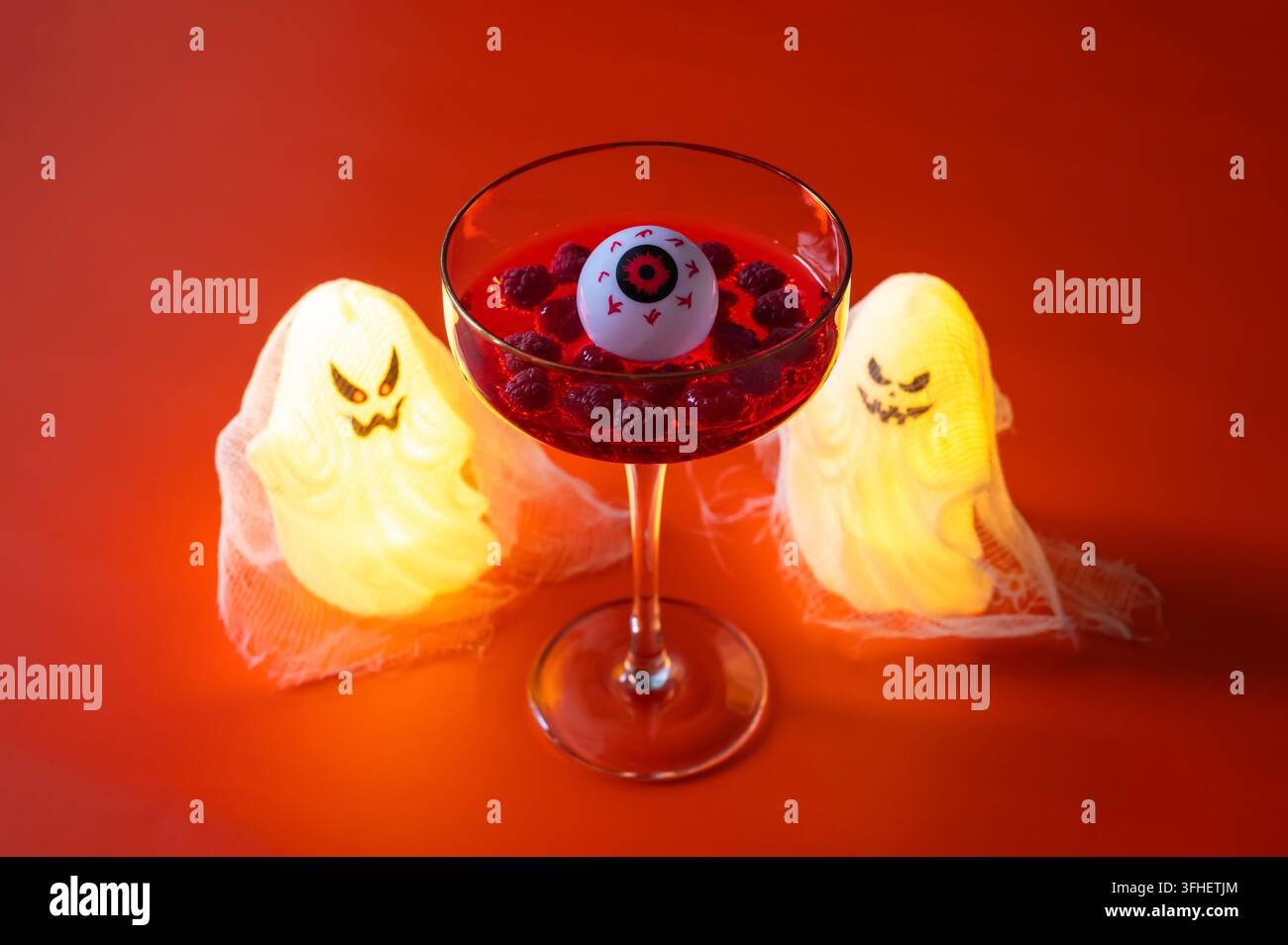 Cocktail d’Halloween avec une garniture d’œil et des baies, flanqué de fantômes lumineux sur un fond orange. Banque D'Images