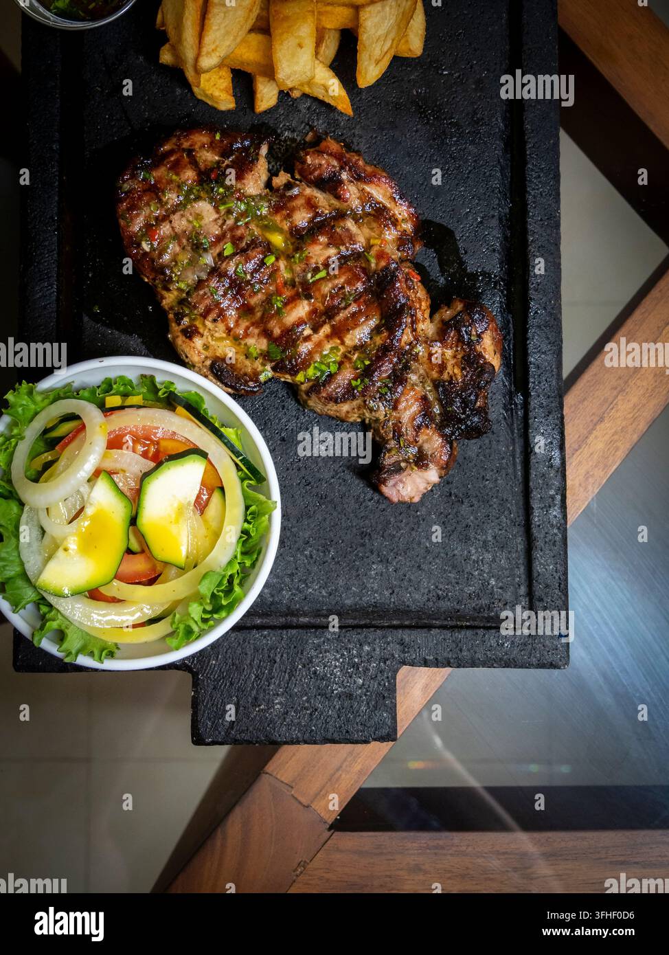 Bondiola de porc grillée servie sur un plateau en pierre noire avec des pommes de terre frites dorées, de la laitue fraîche, une tomate, une salade de courgettes et d'oignons, ainsi qu'une huile aromatisée Banque D'Images