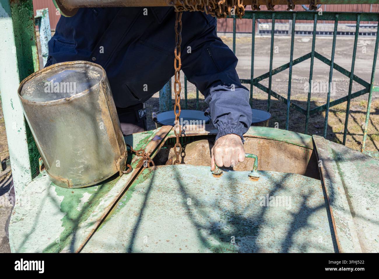 Person ouvrant une couverture en fer usée sur un puits d'eau de jardin dans une cour arrière avec un seau suspendu à proximité, reflétant le travail acharné, l'utilité rurale et le patrimoine Banque D'Images