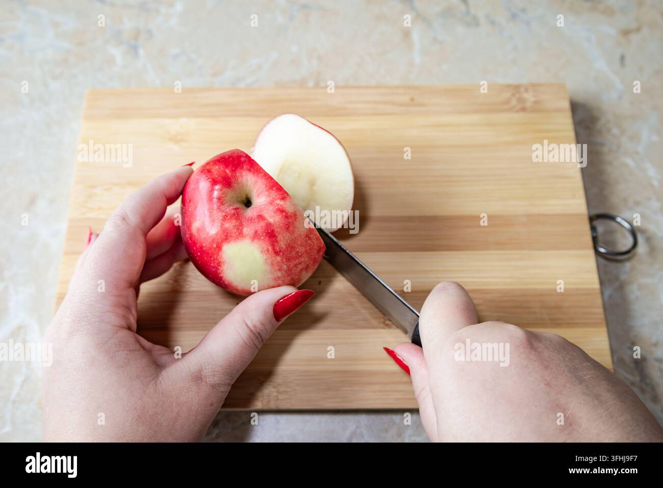 Des mains aux ongles rouges utilisent un couteau de cuisine bien affûté pour couper une pomme rouge fraîche posée sur une planche en bois sur un plan de travail de cuisine. Le plan aérien ca Banque D'Images