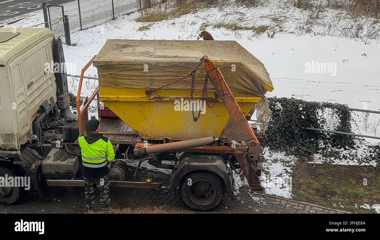 Un opérateur portant un équipement de sécurité réfléchissant fixe une benne à ordures couverte sur une plate-forme de camion hydraulique dans des conditions hivernales, représentant l'efficacité Banque D'Images