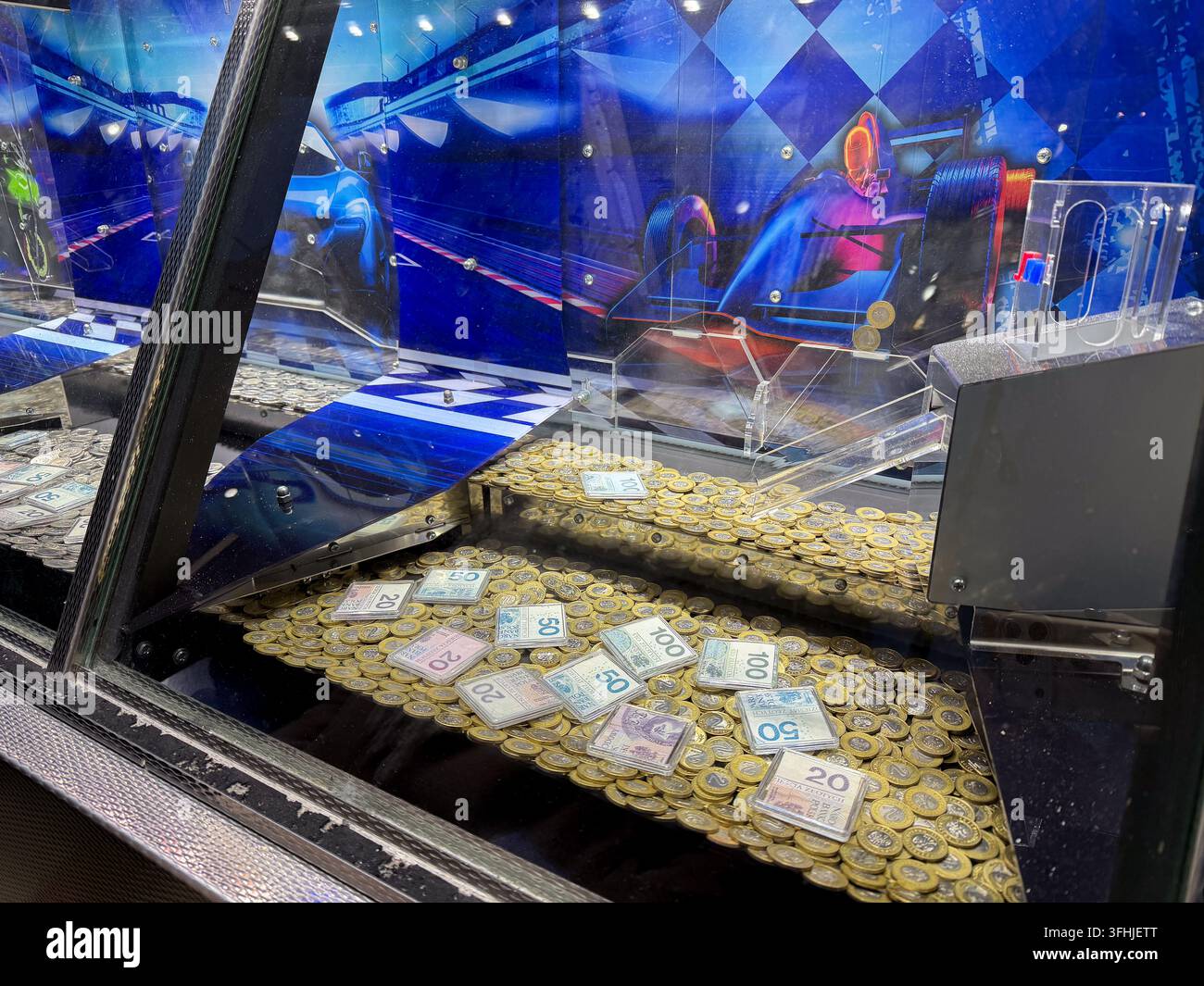 Un jeu d'arcade avec des jetons métalliques et des billets reposant sur une plateforme mobile dans un cabinet lumineux affichant des visuels de sport automobile pour attirer les joueurs Banque D'Images