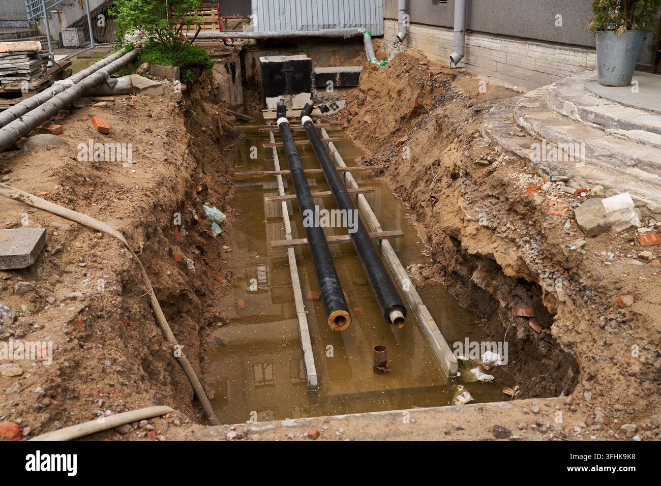 Tranchée de construction pour la rénovation urbaine dans une cour à côté d'un bâtiment avec deux gros tuyaux noirs enjambant une excavation boueuse sur des supports en bois et Banque D'Images