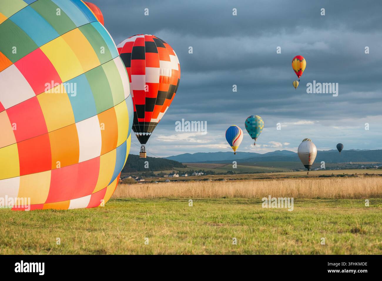 Grand ballon à air chaud coloré à carreaux au premier plan sur le champ avec un ballon rouge qui décolle et des ballons flottants contre un ciel nuageux sur une vaste montagne Banque D'Images
