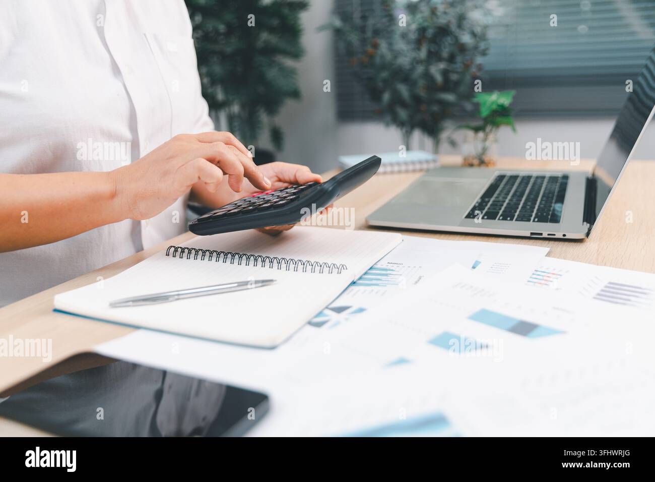 Comptable utilisant une calculatrice au-dessus d'un cahier et de rapports financiers à côté d'un ordinateur portable, représentant la budgétisation, la comptabilité, le suivi des dépenses et la préparation des impôts Banque D'Images