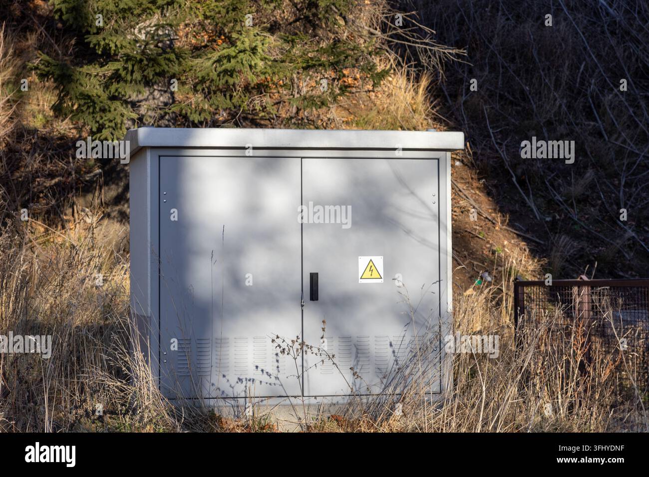 Un cabinet de distribution d'énergie métallique se dresse dans un terrain herbeux sauvage, avec un symbole de haute tension qui illustre le contrôle des infrastructures et l'électricité Banque D'Images