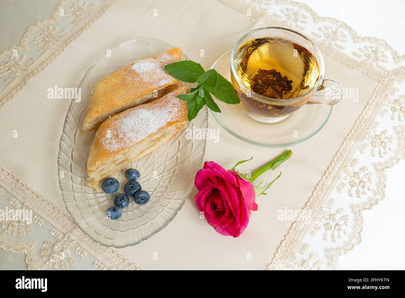 Il y a deux parts de tarte aux pommes, des myrtilles et des feuilles de menthe sur l'assiette, une tasse de thé à côté, et la table est décorée d'une rose vivante. Banque D'Images