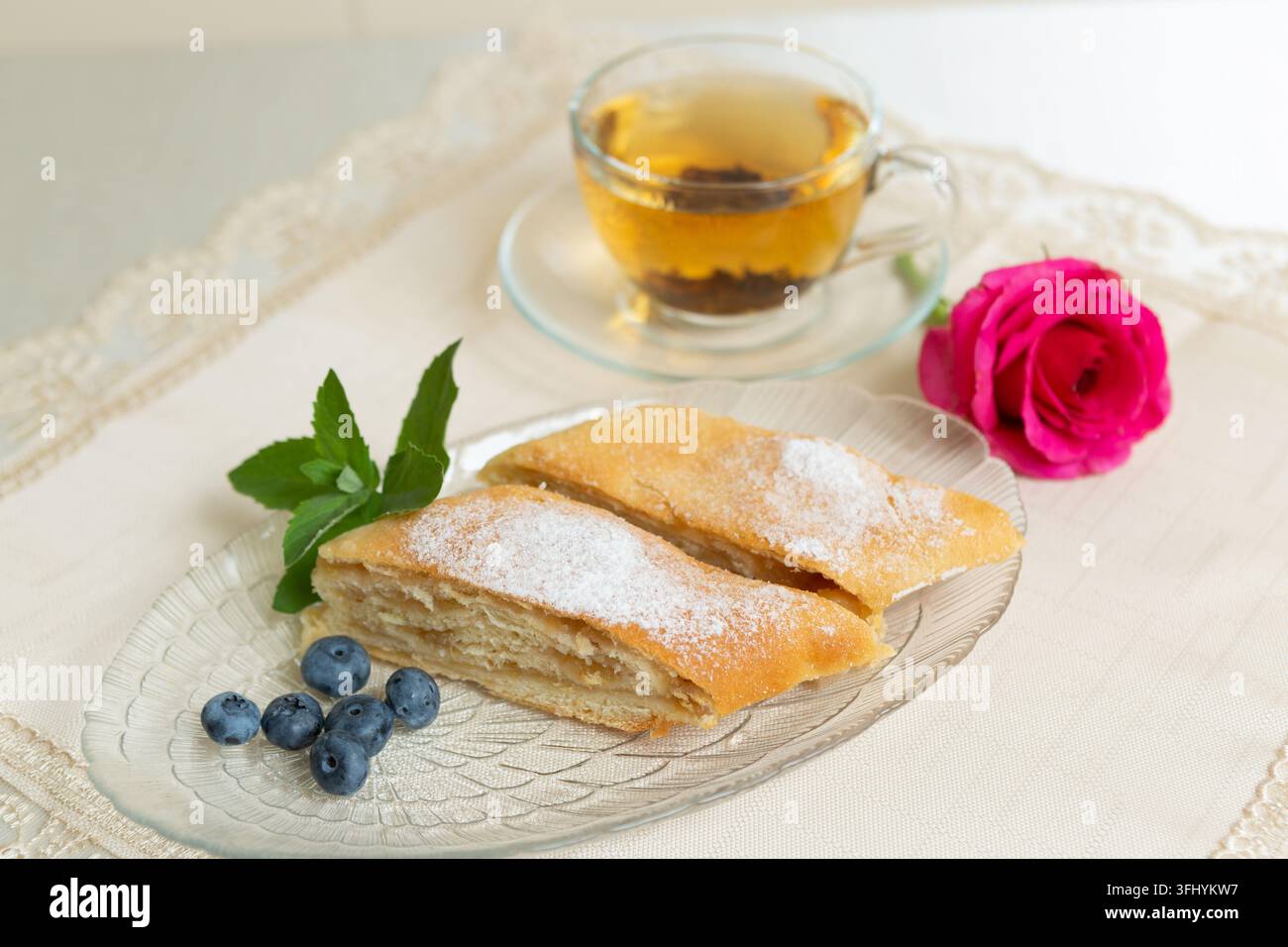 Il y a deux parts de tarte aux pommes, des myrtilles et des feuilles de menthe sur l'assiette, une tasse de thé à côté, et la table est décorée d'une rose vivante. Banque D'Images