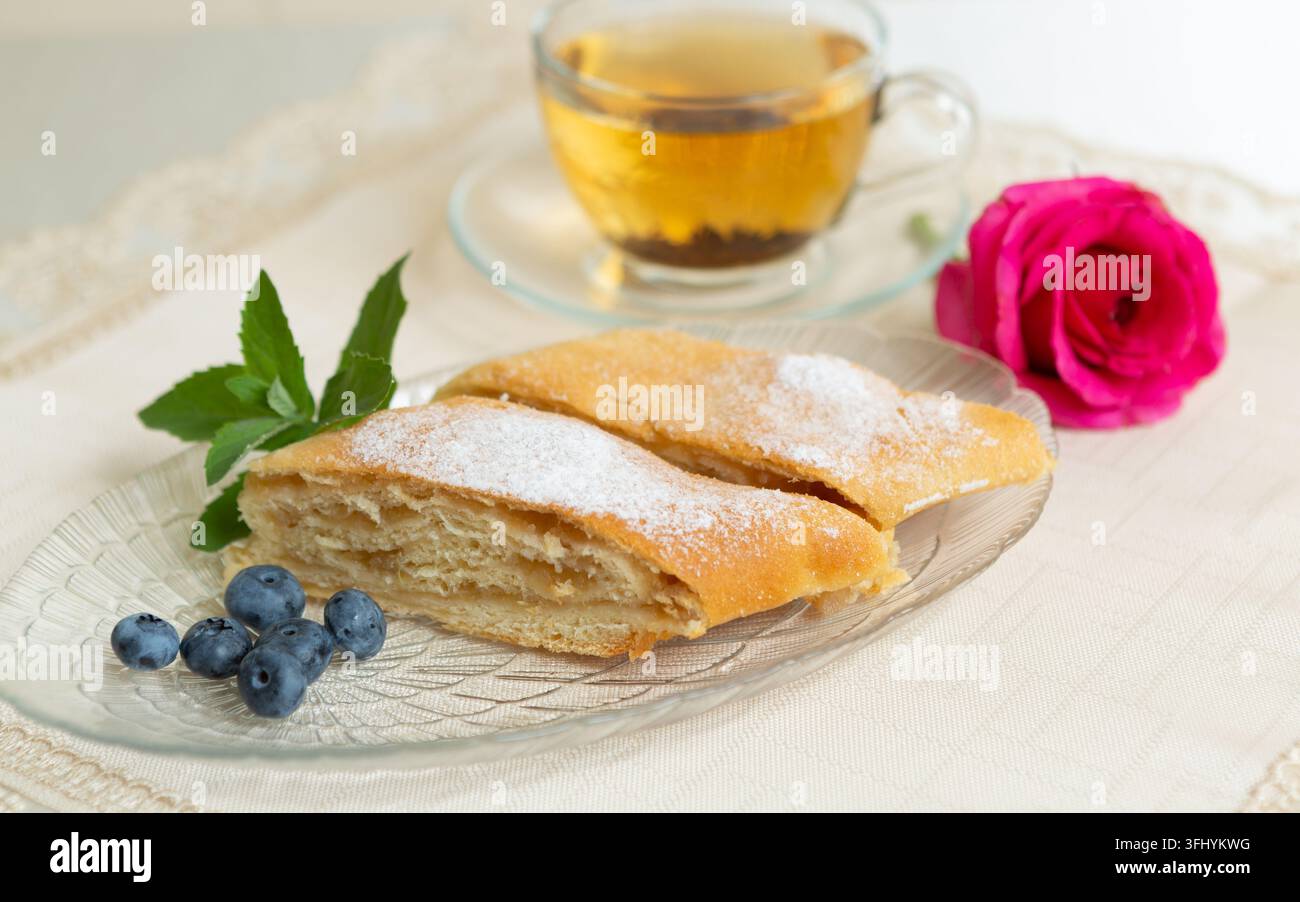 Il y a deux parts de tarte aux pommes, des myrtilles et des feuilles de menthe sur l'assiette, une tasse de thé à côté, et la table est décorée d'une rose vivante. Banque D'Images