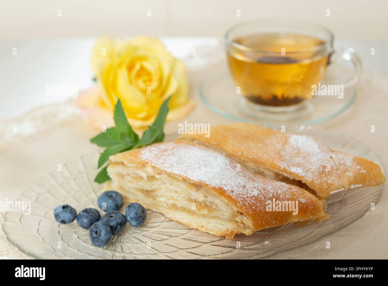 Il y a deux parts de tarte aux pommes, des myrtilles et des feuilles de menthe sur l'assiette, une tasse de thé à côté, et la table est décorée d'une rose vivante. Banque D'Images