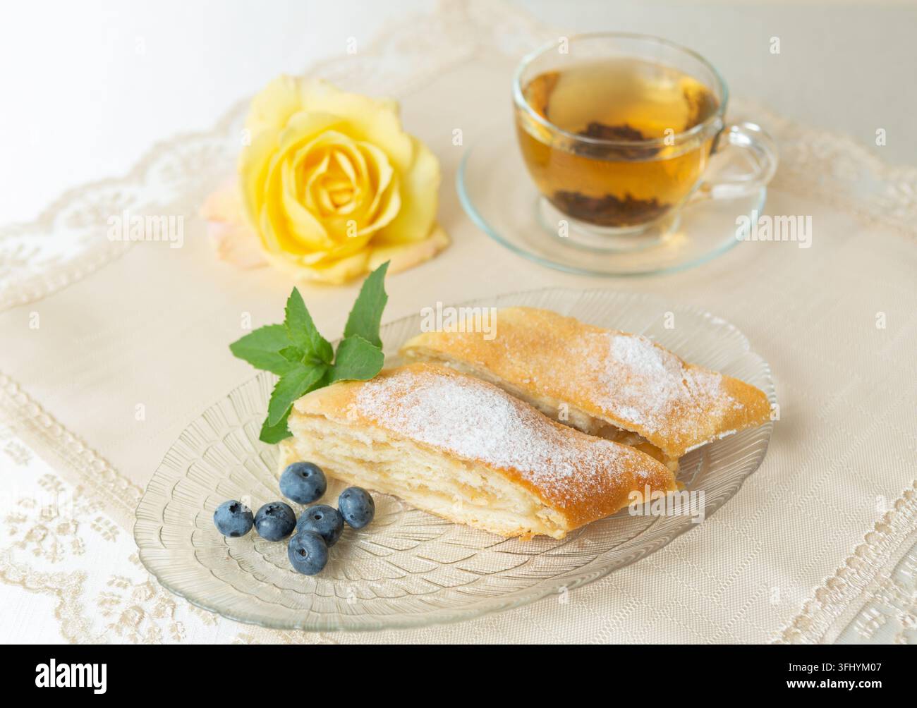 Il y a deux parts de tarte aux pommes, des myrtilles et des feuilles de menthe sur l'assiette, une tasse de thé à côté, et la table est décorée d'une rose vivante. Banque D'Images