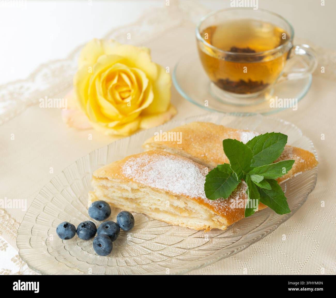 Il y a deux parts de tarte aux pommes, des myrtilles et des feuilles de menthe sur l'assiette, une tasse de thé à côté, et la table est décorée d'une rose vivante. Banque D'Images