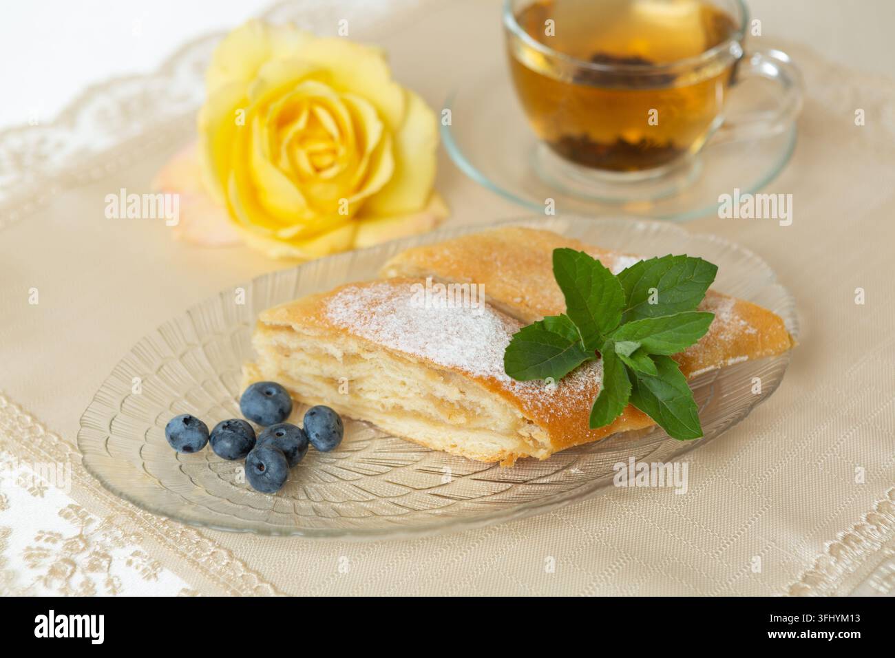 Il y a deux parts de tarte aux pommes, des myrtilles et des feuilles de menthe sur l'assiette, une tasse de thé à côté, et la table est décorée d'une rose vivante. Banque D'Images