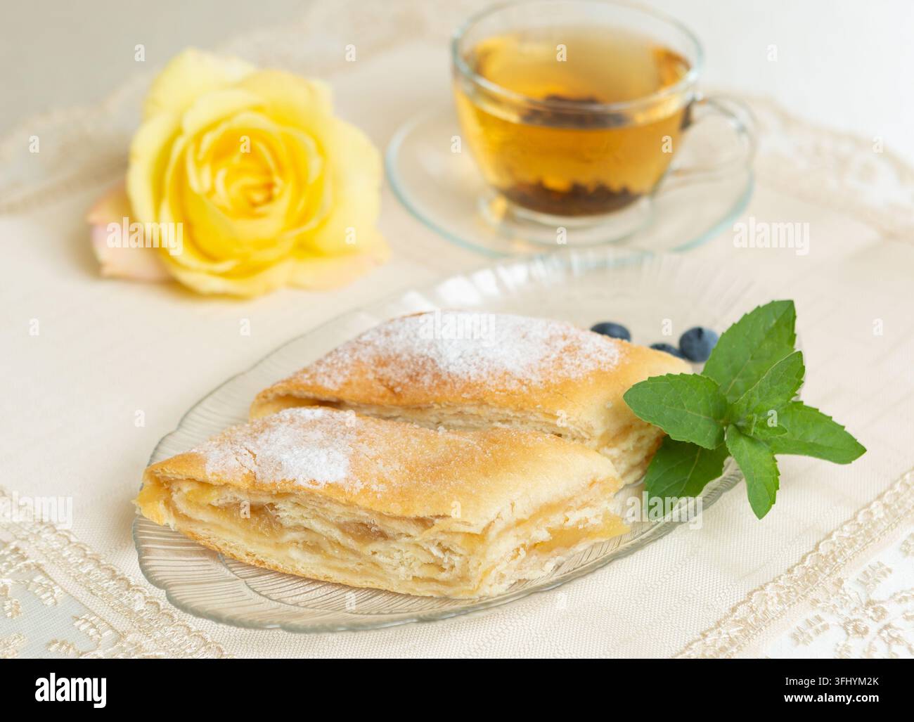 Il y a deux parts de tarte aux pommes, des myrtilles et des feuilles de menthe sur l'assiette, une tasse de thé à côté, et la table est décorée d'une rose vivante Banque D'Images
