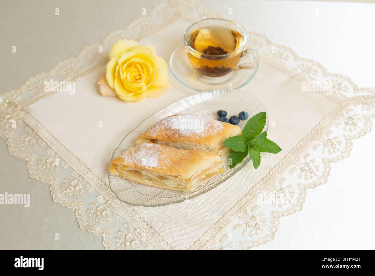 Il y a deux parts de tarte aux pommes, des myrtilles et des feuilles de menthe sur l'assiette, une tasse de thé à côté, et la table est décorée d'une rose vivante Banque D'Images