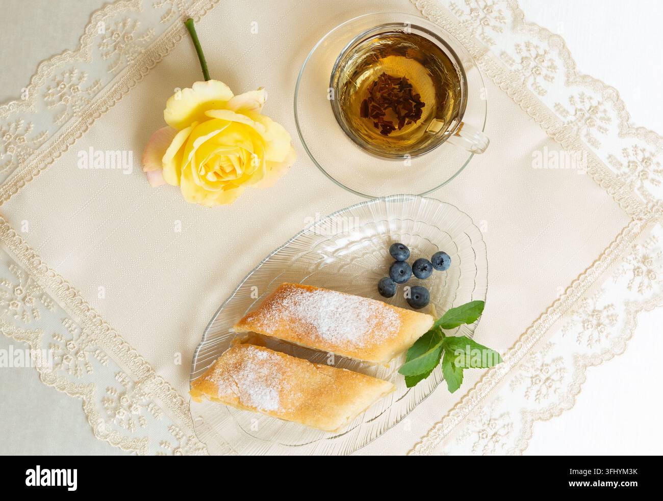 Il y a deux parts de tarte aux pommes, des myrtilles et des feuilles de menthe sur l'assiette, une tasse de thé à côté, et la table est décorée d'une rose vivante Banque D'Images