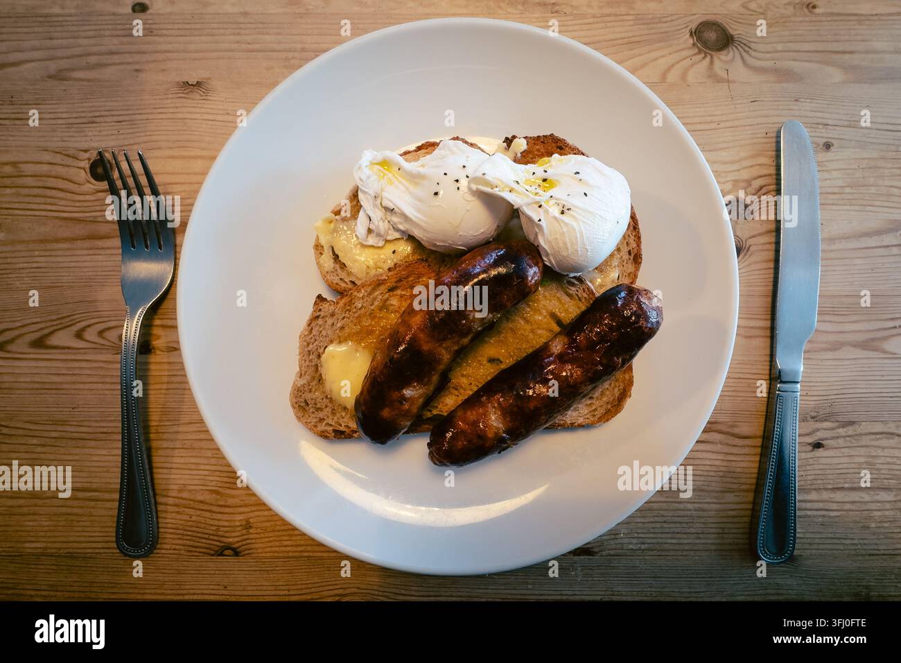 Vue aérienne d'une assiette de petit-déjeuner composée de saucisses grillées, d'œufs pochés et de toasts beurrés sur une table en bois. - Image de stock capturée avec un smartphone