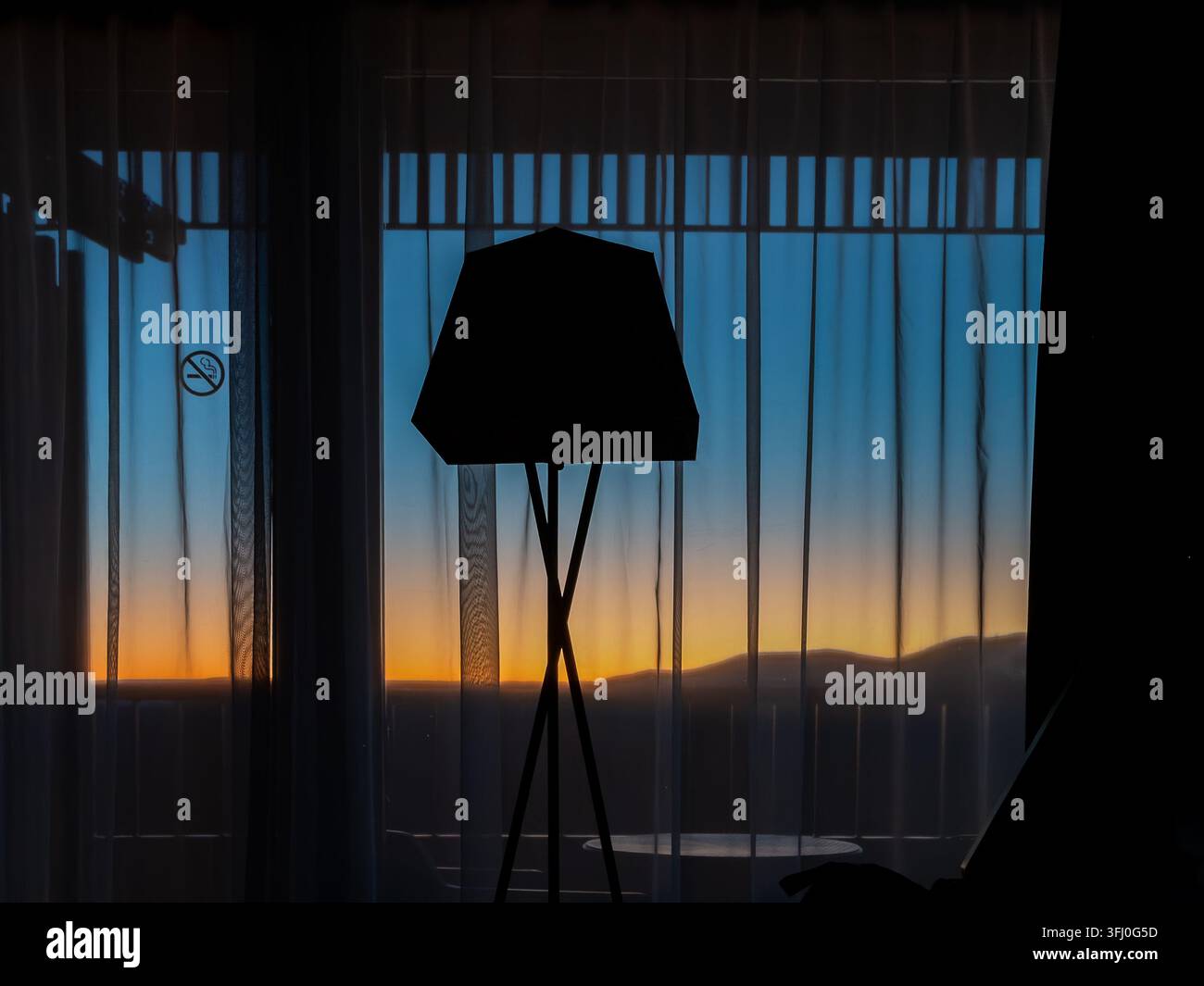 La silhouette d’une lampe trépied encadre un fond de coucher de soleil orange lumineux à travers des rideaux de fenêtre transparents, créant une composition intérieure minimaliste qui Banque D'Images